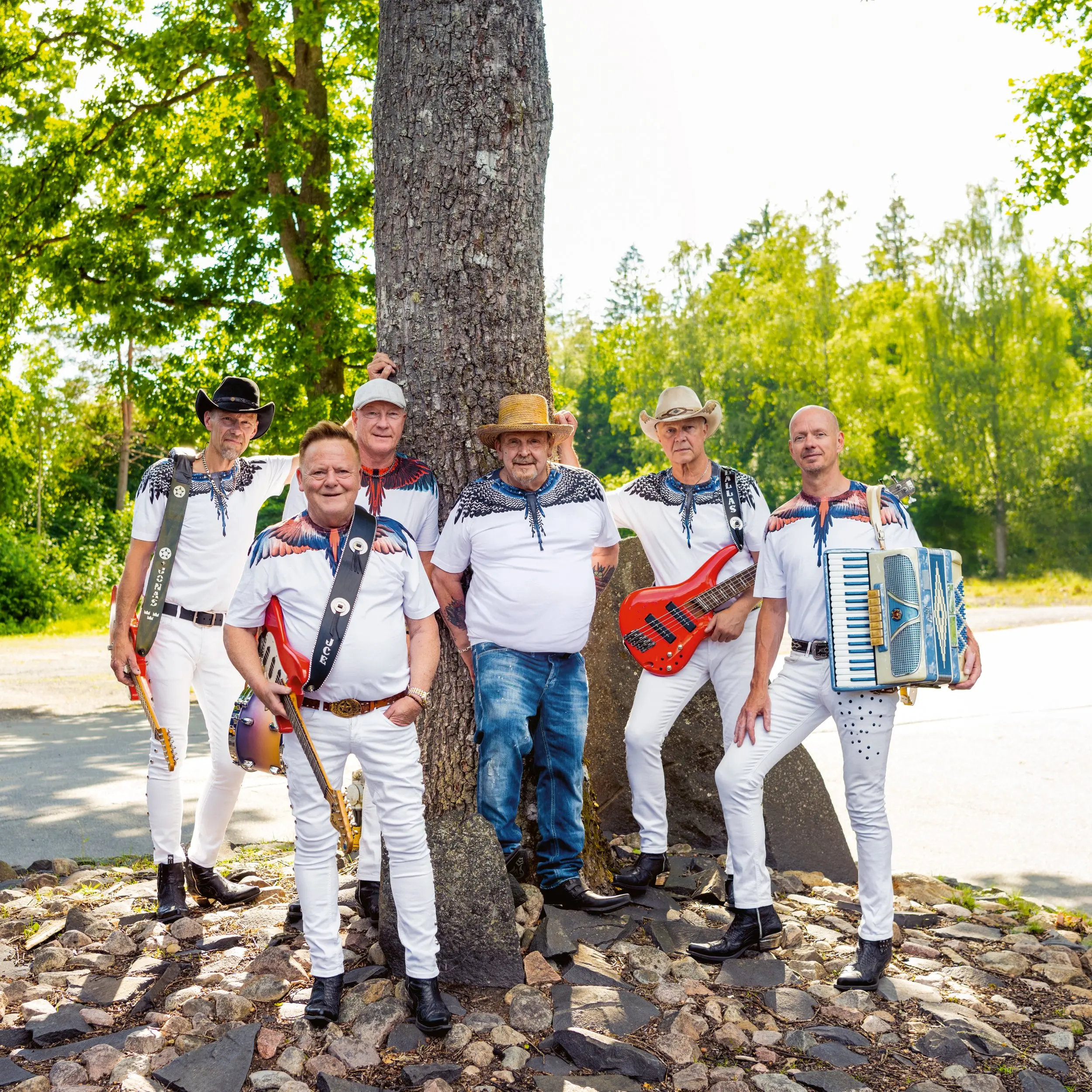 En grupp på sex män i matchande kläder poserar utomhus med musikinstrument, inklusive gitarrer och ett dragspel, vid ett träd.